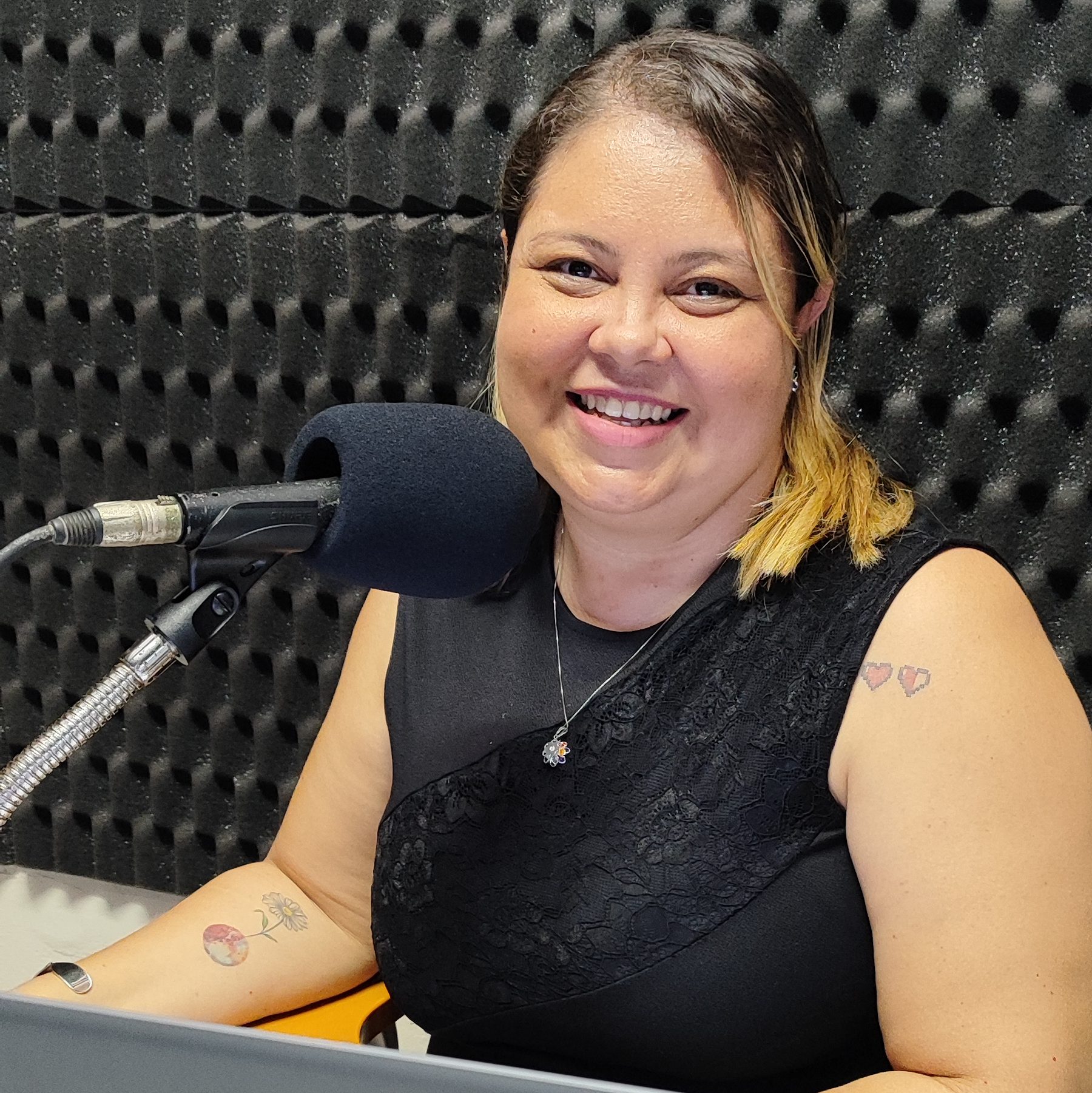 Foto de Dayse Martins. Mulher branca e cabelos loiros. Ela usa um vestido preto. À sua direita um microfone.