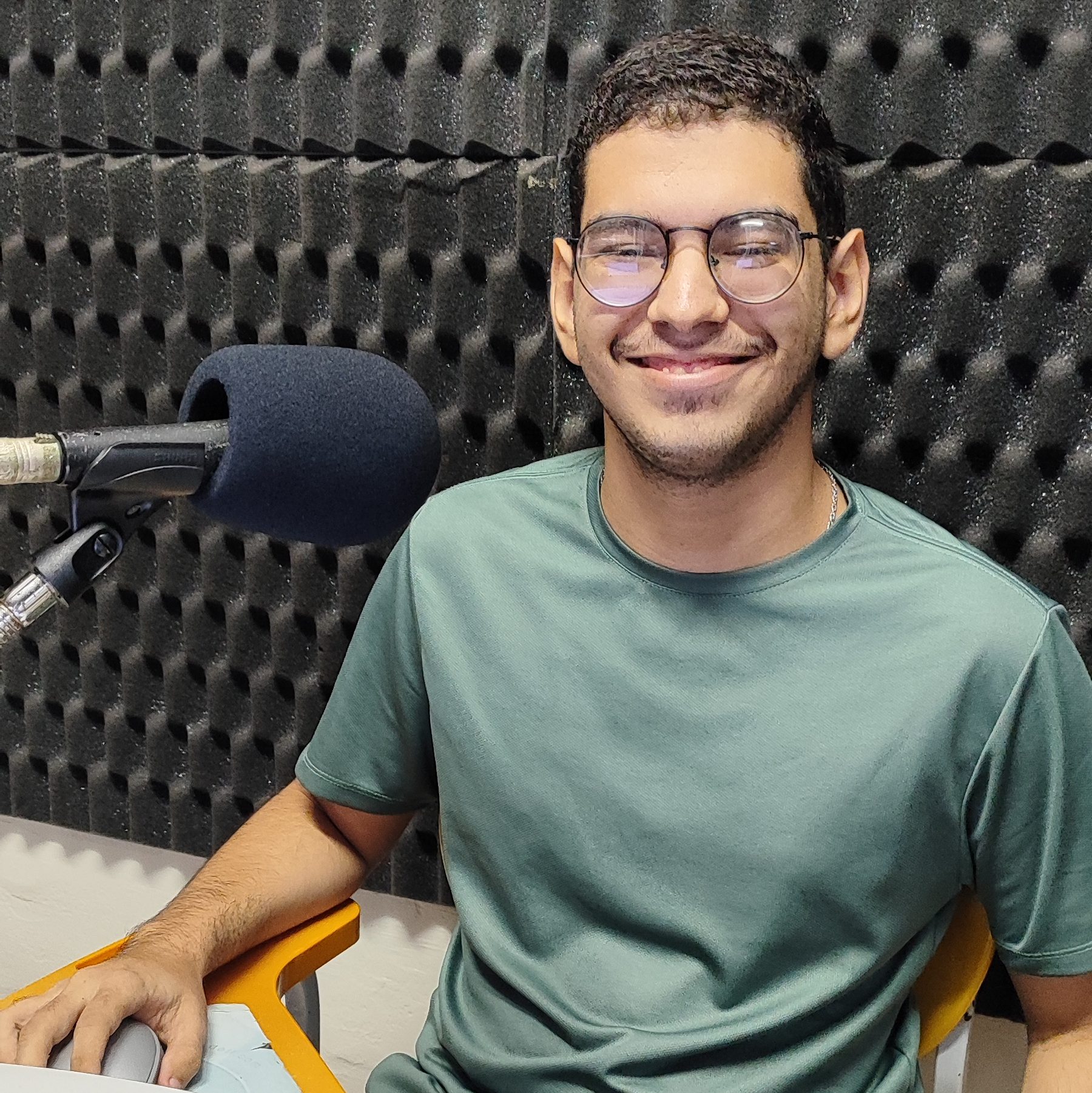 Foto de Guilherme Santana. Jovem branco de cabelos pretos curtos e óculos. Veste camisa verde. À sua direita um microfone. Ele sorri.