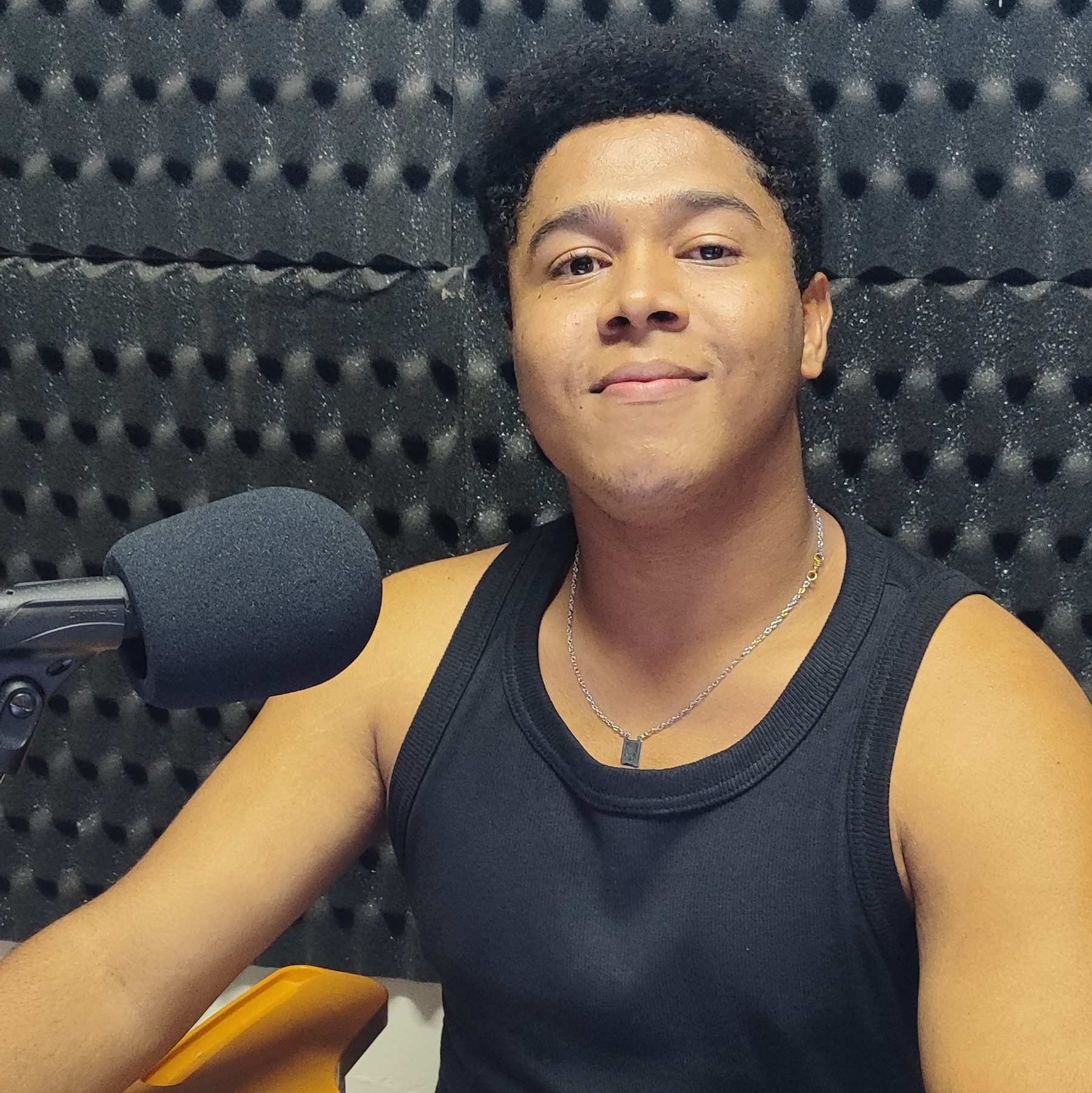 Foto de Marcelo Reis. Jovem negro de pele retinta. Cabelo preto curto. Usa camisa regata preta e um cordão de metal no pescoço. À sua direita um microfone.