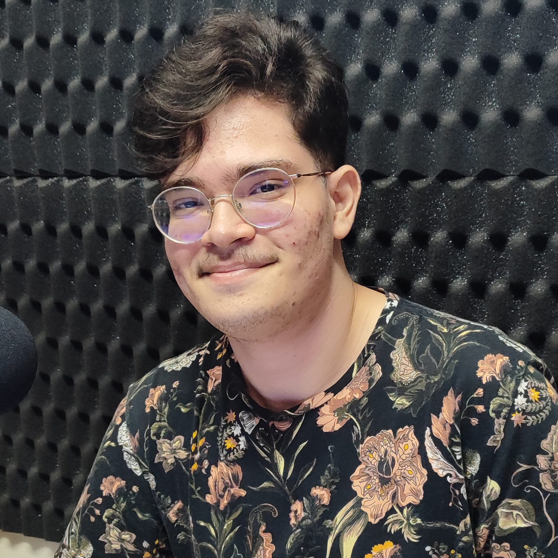 Foto de Raphael Fontoura. Jovem branco de cabelos pretos, lisos e curtos. Usa óculos e uma camisa preta com estampas de flores. Ele sorri.