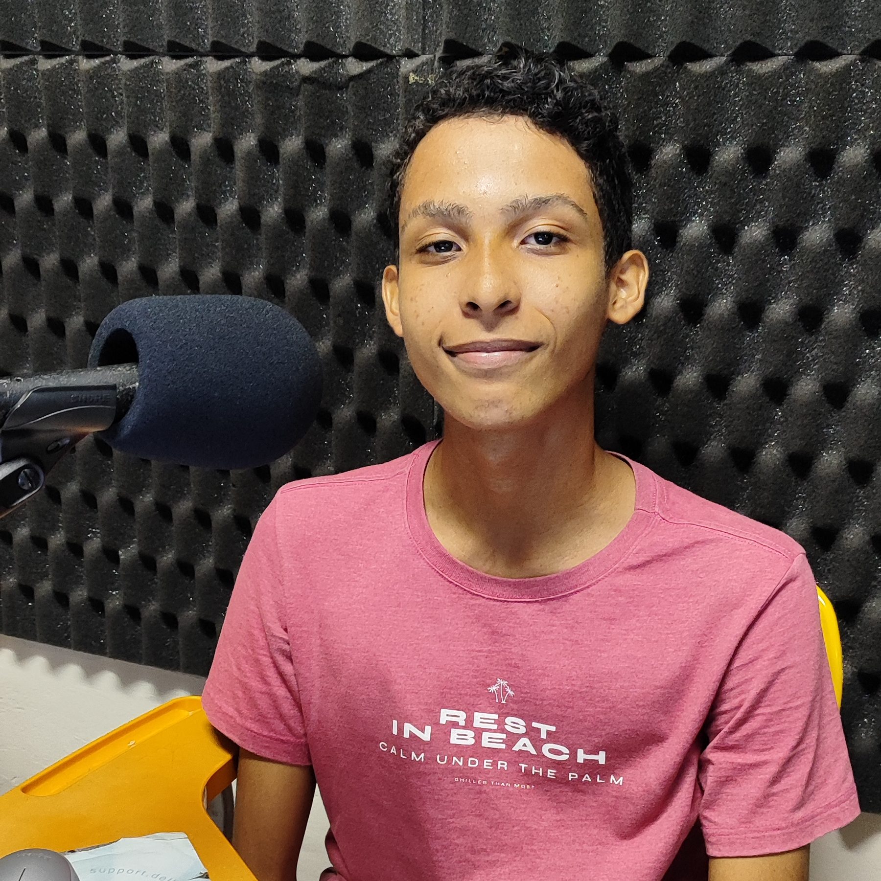 Foto de Ricardo Miranda. Jovem negro de pelo não retinta e cabelos pretos curtos. Usa uma camisa em tom de rosa. À sua direita um microfone.