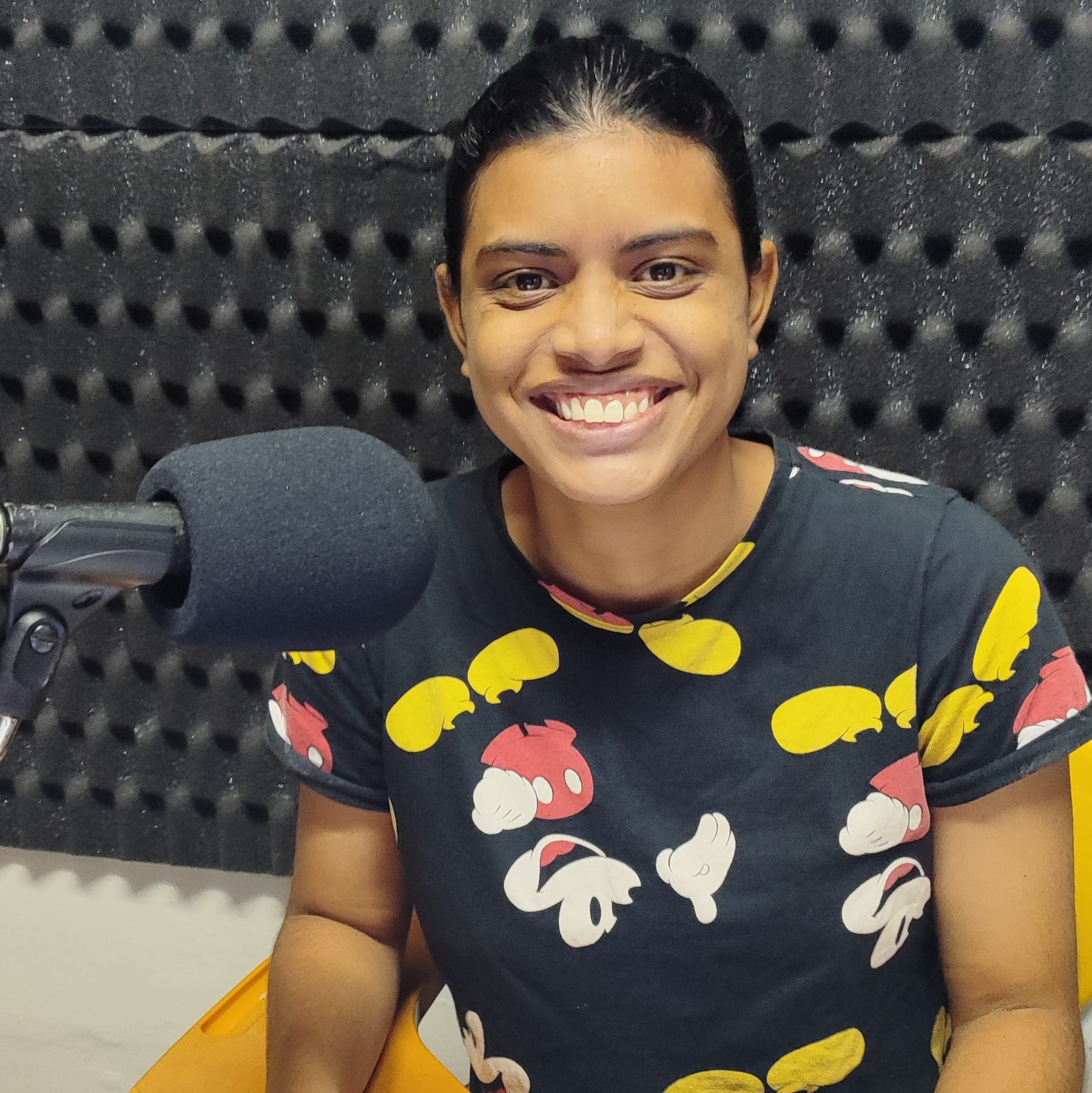 Foto de Yasmin Borges. Jovem negra de pele retinta e cabelos pretos lisos. Ela usa uma camisa preta com estampas do Mickey Mouse. À sua direita um microfone. Yasmin sorri.